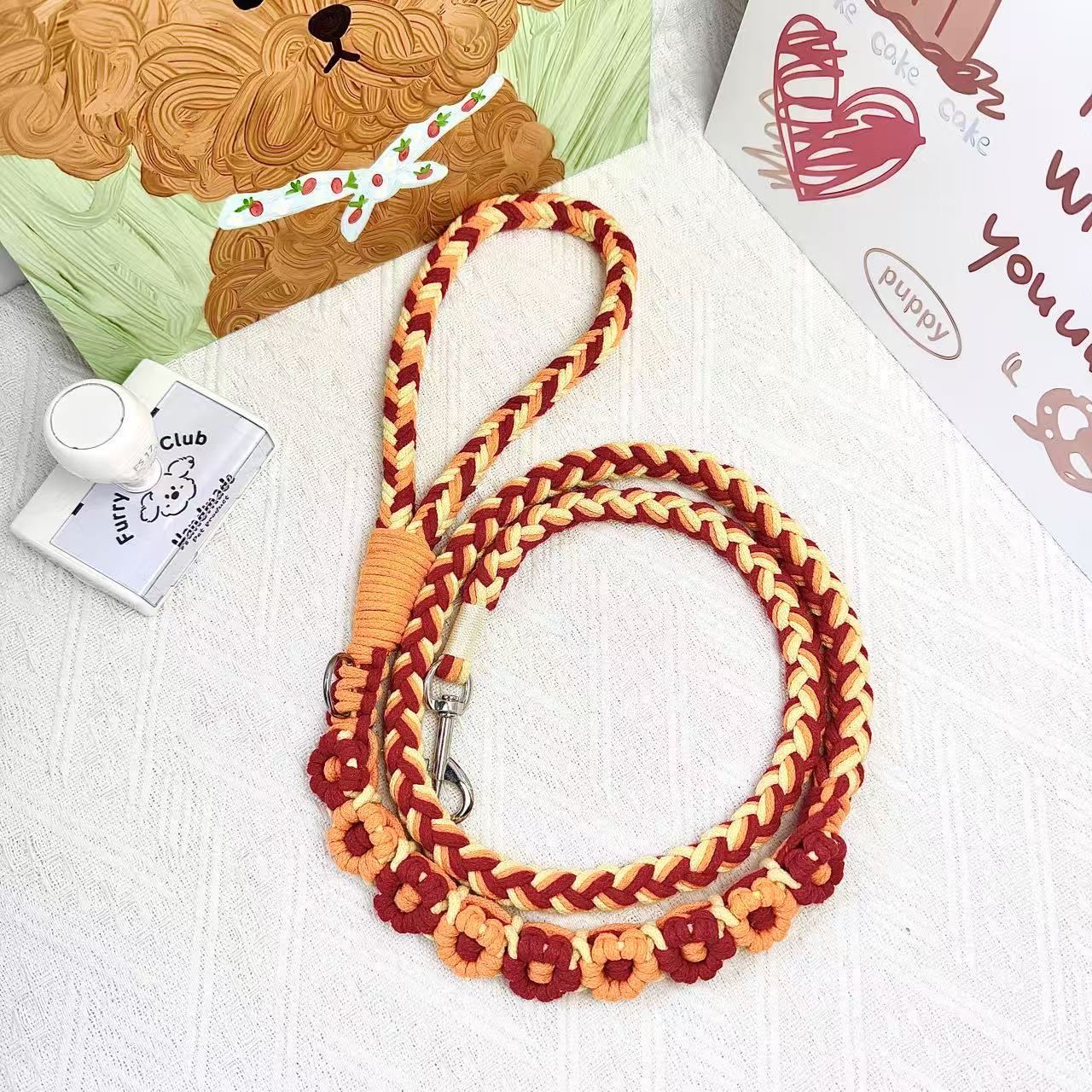 A handmade dog leash in orange and red with floral accents, featuring a metal clasp, placed on a textured white fabric background.