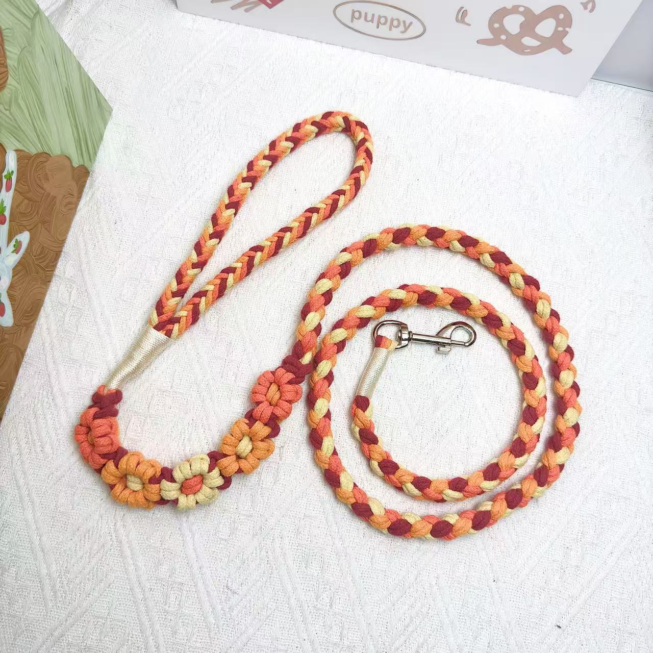 A handmade dog leash in orange and yellow with floral accents, featuring a metal clasp, placed on a textured white fabric background.