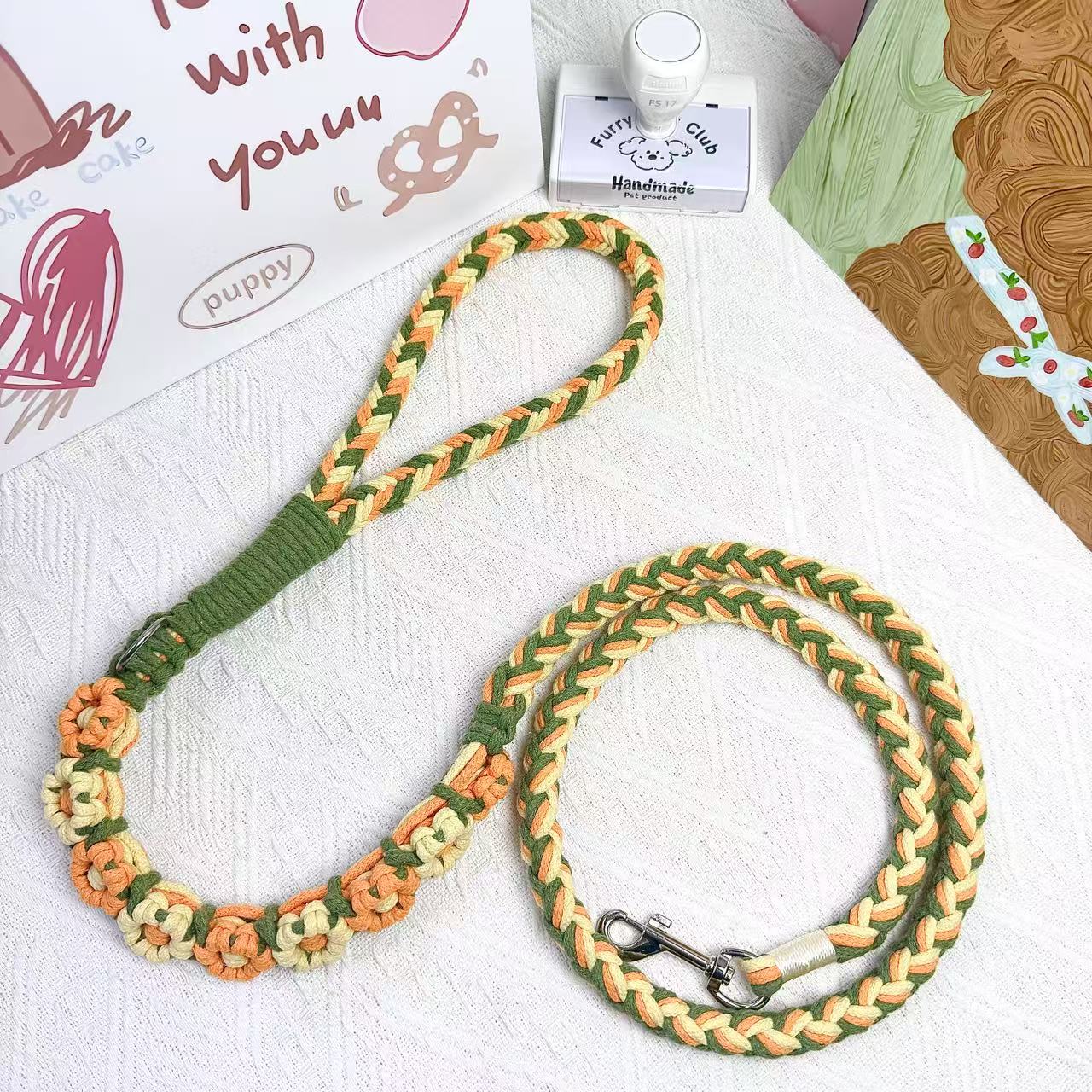 A handmade dog leash in orange and yellow with floral accents, featuring a metal clasp, placed on a textured white fabric background.