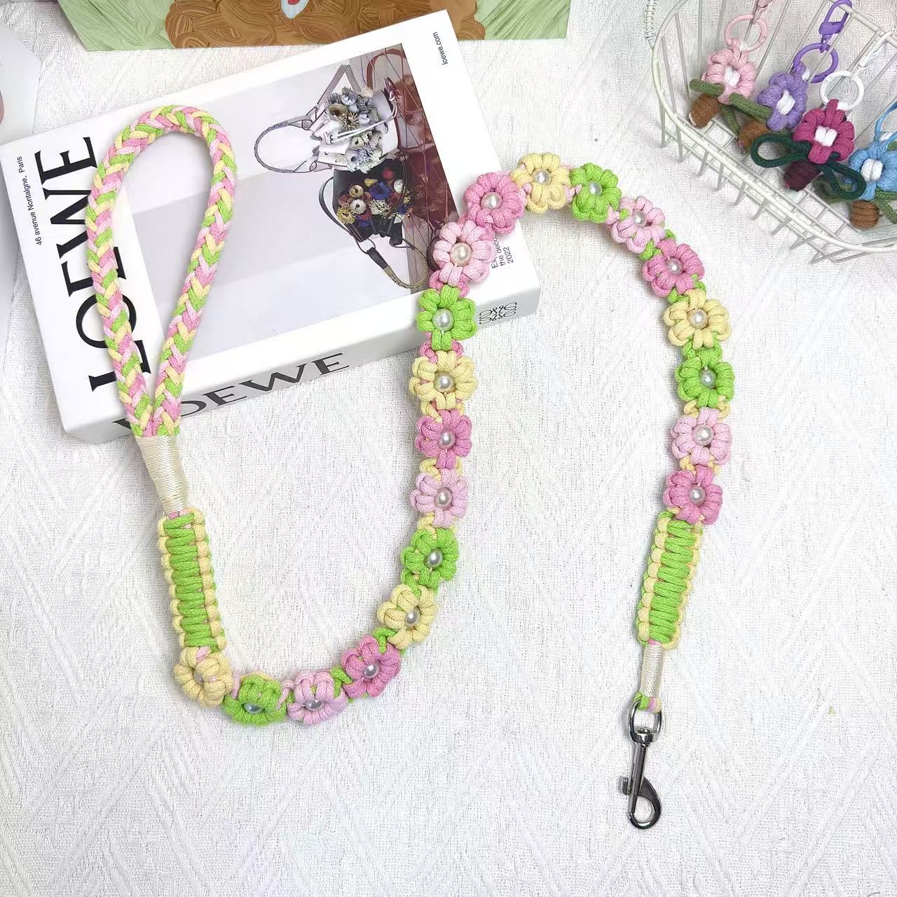 A handmade dog leash in pink and green with floral accents and pearl details, featuring a metal clasp, placed on a textured white fabric background.