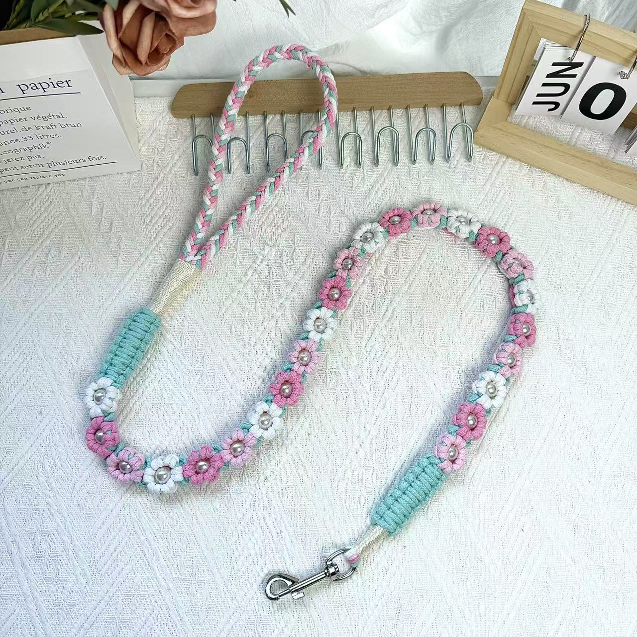 A handmade dog leash in pink and white with floral accents and pearl details, featuring a metal clasp, placed on a textured white fabric background.