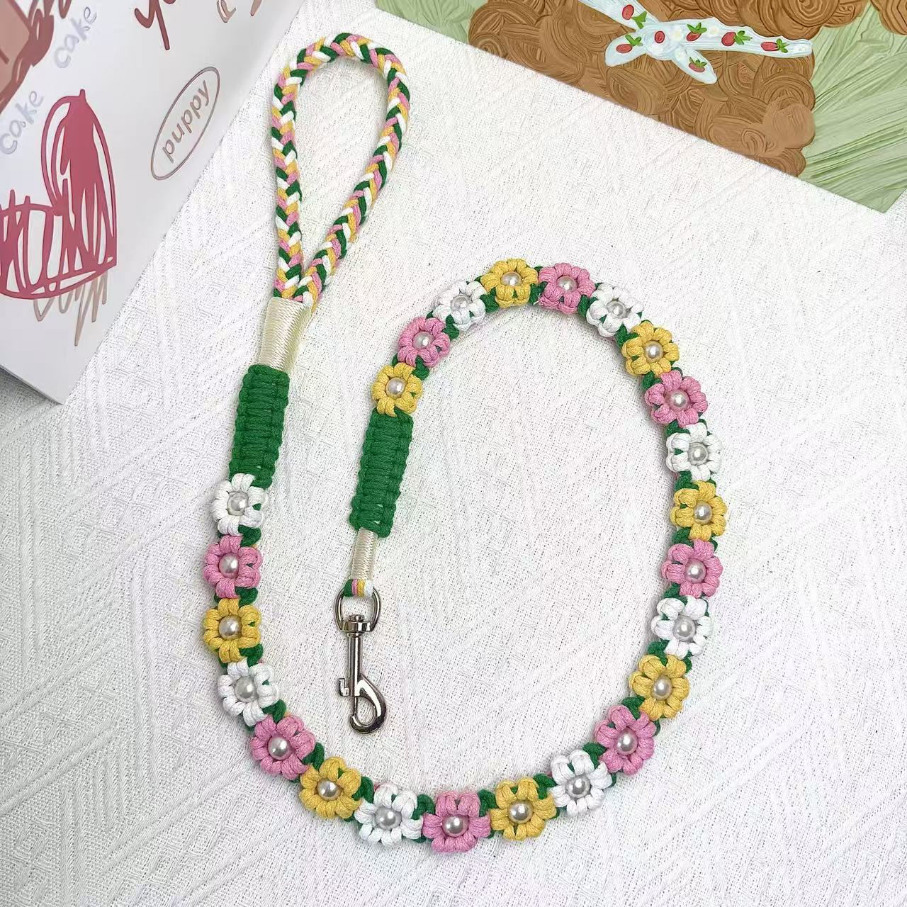 A handmade dog leash in pink and yellow with floral accents and pearl details, featuring a metal clasp, placed on a textured white fabric background.