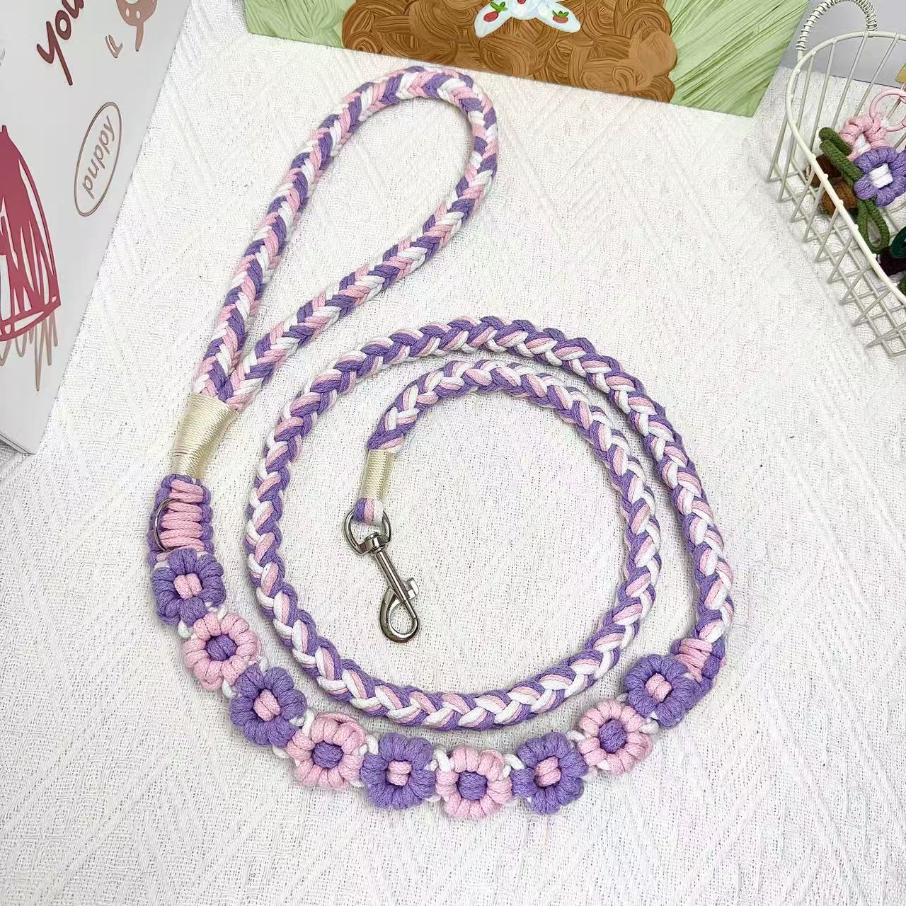 A handmade dog leash in pink and purple with floral accents, featuring a metal clasp, placed on a textured white fabric background.