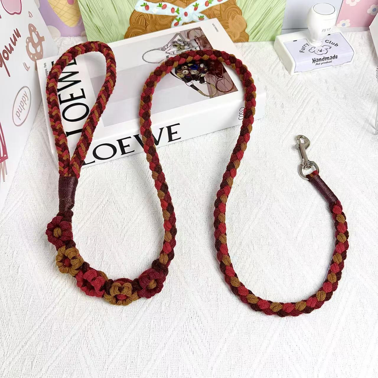A handmade dog leash in red and brown with floral accents, featuring a metal clasp, placed on a textured white fabric background.