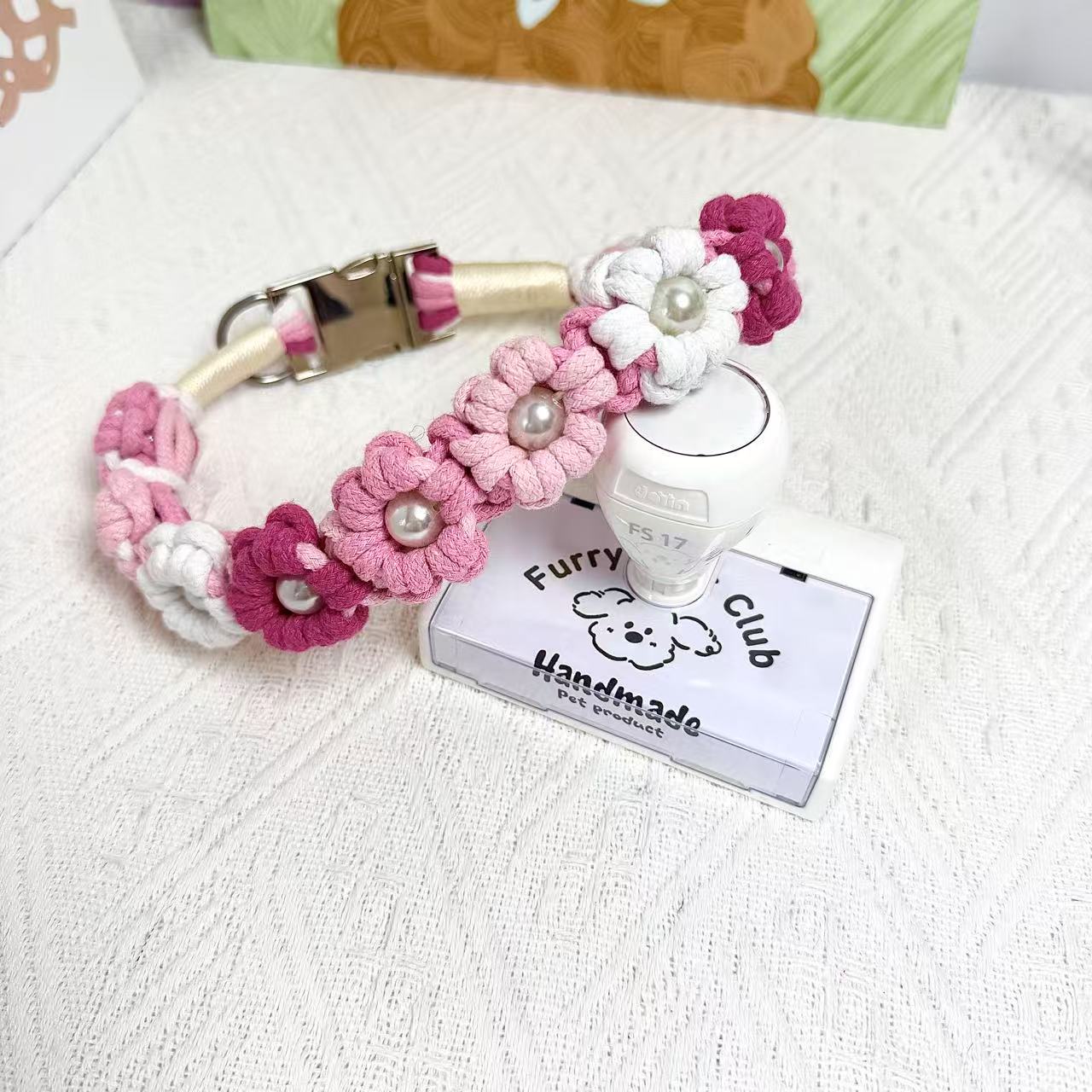 A handmade dog collar in pink and red with floral accents and pearl details, featuring a metal buckle, placed on a textured white fabric background.