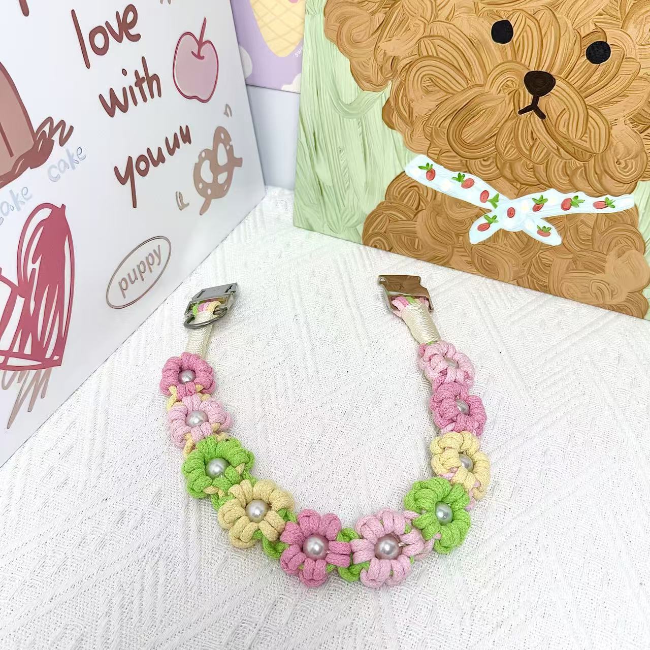 A handmade dog collar in pink, green, and yellow with floral accents and pearl details, featuring a metal buckle, placed on a textured white fabric background.
