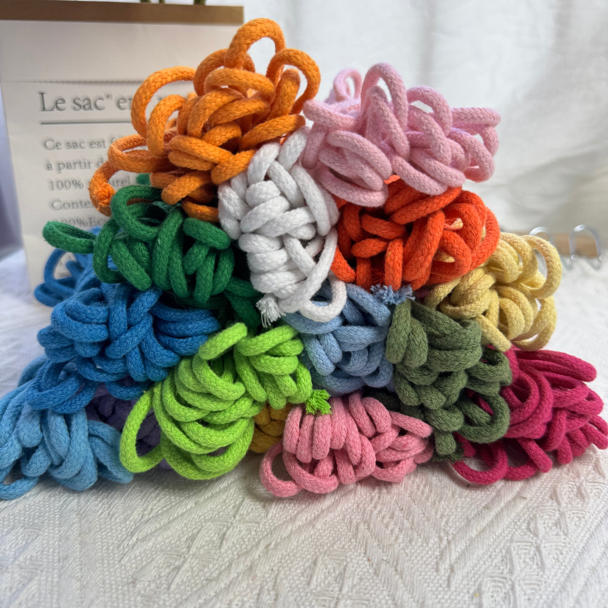 A colorful assortment of 4mm cotton ropes stacked in a pyramid shape, featuring vibrant hues like orange, pink, green, blue, and white, displayed on a textured fabric background.