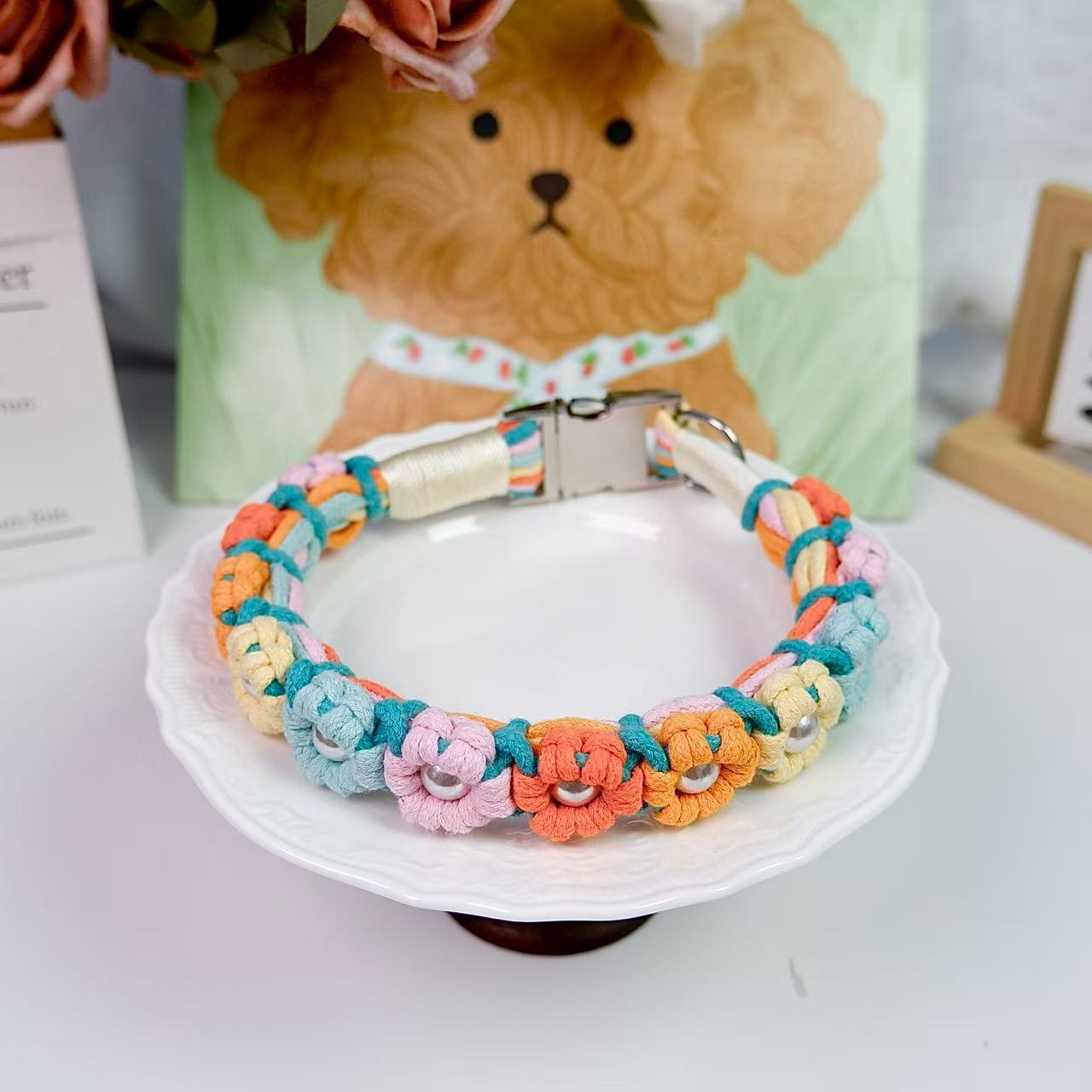 Handwoven dog collar with colorful flowers and pearl accents, featuring a stainless steel clasp and D-ring, on a white dish.