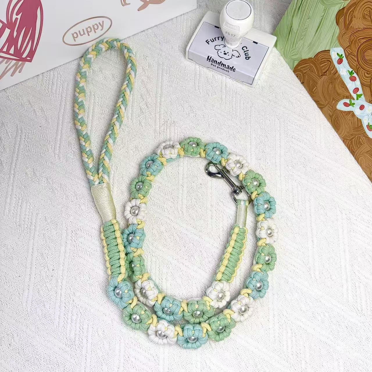 A handmade dog leash in white and green with floral accents and pearl details, featuring a metal clasp, placed on a textured white fabric background.